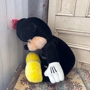 May include: A plush black and white character toy with yellow feet and a red polka dot bow. The toy has white gloves with three black lines on the palm. The toy is sitting on a patterned rug.