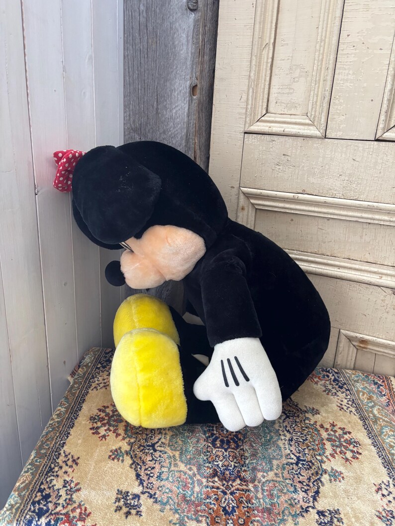 May include: A plush black and white character toy with yellow feet and a red polka dot bow. The toy has white gloves with three black lines on the palm. The toy is sitting on a patterned rug.