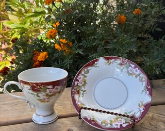 May include: A vintage teacup and saucer set. The teacup has a burgundy rim and floral design, with a white interior and gold trim. The matching saucer features a similar floral pattern and burgundy border. The set is displayed on a wooden surface.