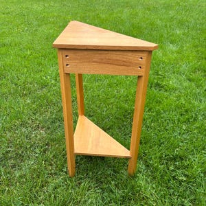 May include: A light brown wooden corner table with a triangular top and a matching triangular lower shelf. The table features three straight legs and a front apron with four visible screw holes, showcasing a natural wood grain finish.