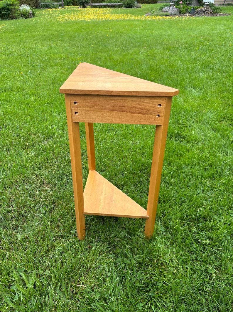 May include: A light brown wooden corner table with a triangular top and a matching triangular lower shelf. The table features three straight legs and a front apron with four visible screw holes, showcasing a natural wood grain finish.