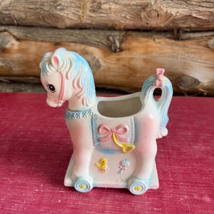 Könnte beinhalten: Ein Vintage-Pflanzgefäß in Form eines Schaukelpferds aus Keramik in Pastellrosa, Blau und Weiß. Das Pferd hat eine rosa Schleife, blaue Mähne und Schweif sowie dekorative Akzente. Der Pflanztopf hat eine rechteckige Basis mit Rädern und ist mit einer gelben Ente und einer Babyrassel verziert.