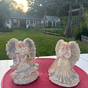 Puede incluir: Dos figuras de ángeles con alas detalladas y vestidos fluidos. Los ángeles tienen una paleta de colores crema y beige, con acentos de azul y dorado. Se colocan sobre un mantel individual circular rojo, con una casa y jardín al fondo.