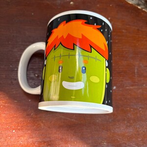 May include: A white ceramic mug featuring a graphic illustration of a green-skinned character with bright orange hair, a stitched forehead, small blue eyes, and a smiling mouth, set against a black background with small white dots.