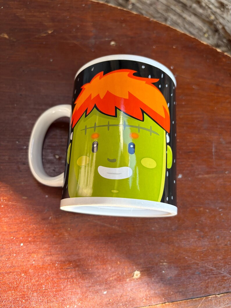 May include: A white ceramic mug featuring a graphic illustration of a green-skinned character with bright orange hair, a stitched forehead, small blue eyes, and a smiling mouth, set against a black background with small white dots.