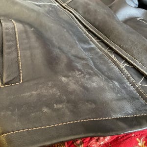 May include: Close-up of a black leather jacket with white stitching. The jacket has a pocket on the left side and a zipper closure. The leather has a worn and distressed look.