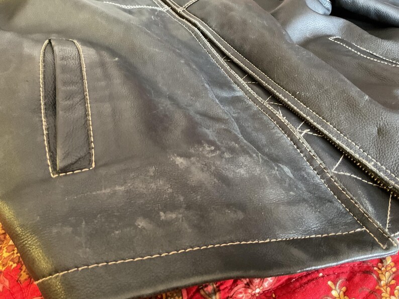 May include: Close-up of a black leather jacket with white stitching. The jacket has a pocket on the left side and a zipper closure. The leather has a worn and distressed look.