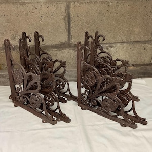 May include: Set of four ornate cast iron shelf brackets with intricate floral designs. The brackets are a dark brown color and have a distressed finish.
