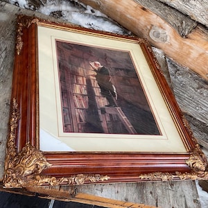 May include: A framed print of a painting depicting a man standing on a ladder in a library, reaching for a book. The frame is made of dark wood with gold accents.