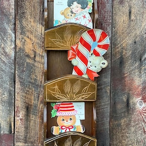 Puede incluir: Un portacartas de pared de madera con tres ranuras. Cada ranura tiene un diseño tallado y contiene una tarjeta de Navidad vintage con un niño, un bastón de caramelo y un hombre de jengibre. Las tarjetas tienen el texto "We Sister! Merry Christmas" y "Have Fun at Christmas!"
