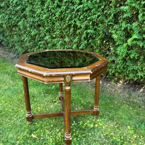 Könnte beinhalten: Ein achteckiger Beistelltisch mit dunkler Glasplatte und Holzrahmen. Der Tisch hat vier kannelierte Beine mit dekorativen goldenen Akzenten. Der Tisch steht auf einem Rasen vor einer grünen Hecke.