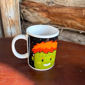 May include: A white ceramic mug featuring a graphic illustration of a green-skinned character with bright orange hair, a stitched forehead scar, and a friendly smiling expression set against a black background with small white speckles.