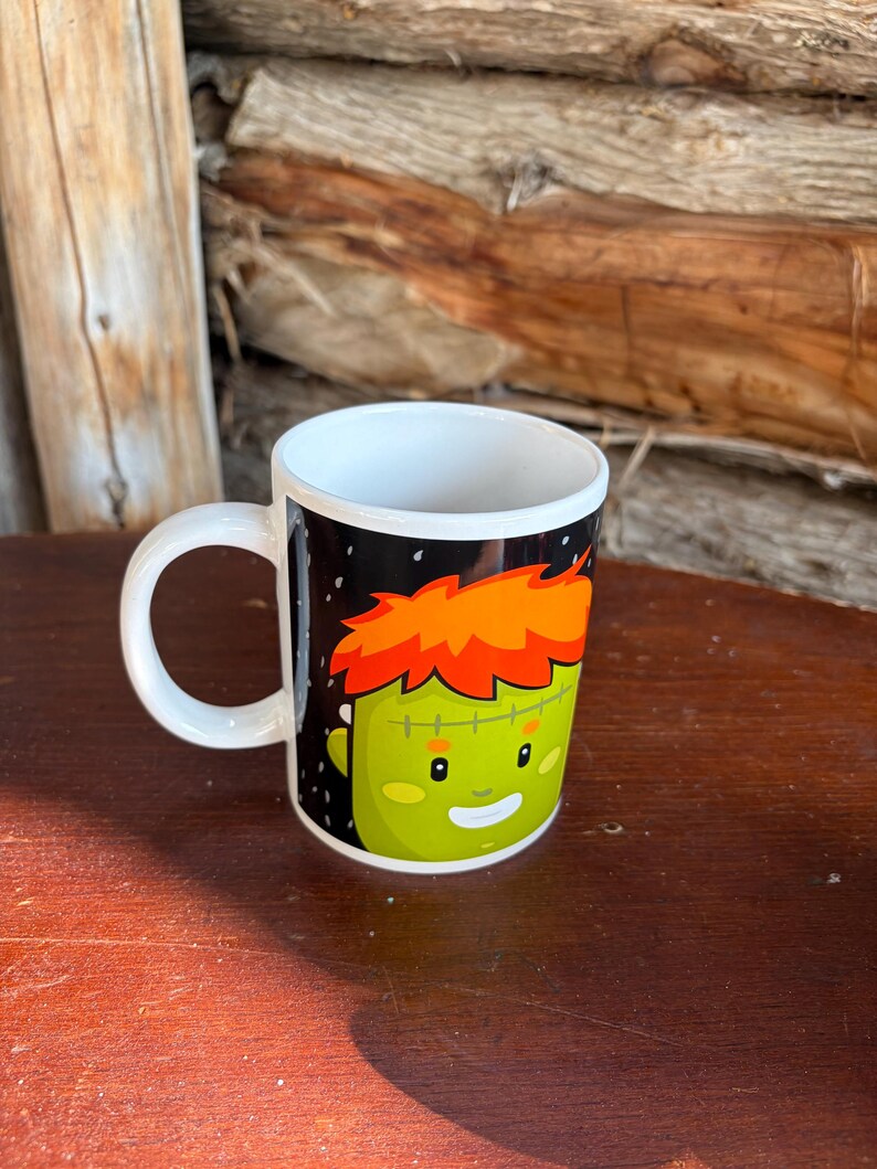 May include: A white ceramic mug featuring a graphic illustration of a green-skinned character with bright orange hair, a stitched forehead scar, and a friendly smiling expression set against a black background with small white speckles.