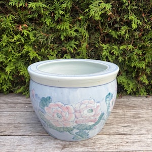 May include: A round ceramic planter with a light blue base colour, featuring hand-painted pink peony flowers and green leaves. The rim is decorated with a blue geometric pattern, and the interior of the bowl is a smooth, pale off-white finish.