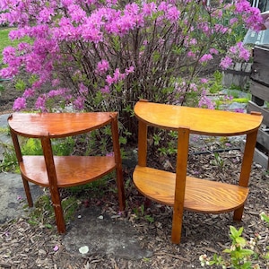 May include: Two wooden side tables with a half-circle design. Each table has two shelves and four legs. The tables are stained in shades of brown and are set outdoors with a backdrop of pink flowering bushes.
