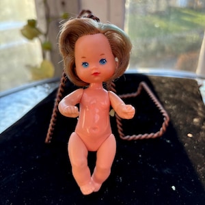 May include: A small plastic doll with brown and blue hair, blue eyes, and a pink body. The doll is wearing a necklace and is sitting on a black surface.