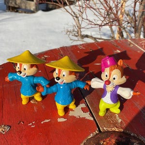 Puede incluir: Tres figuras de plástico de Chip y Dale en diferentes disfraces. Uno lleva un fez rojo, otro lleva un sombrero amarillo y el tercero lleva un sombrero amarillo. Los tres llevan una camisa azul y pantalones amarillos.
