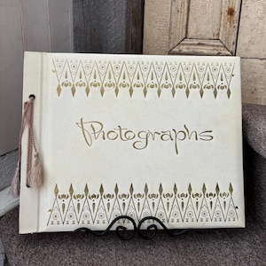 May include: Cream-coloured photo album with gold decorative accents and the word "Photographs" in elegant script. The album features a tassel and is displayed on a black metal stand. The cover has a repeating geometric pattern.