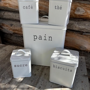 Peut inclure: Cinq boîtes en métal blanc avec des inscriptions noires. Les boîtes sont étiquetées "café", "thé", "pain", "sucre" et "biscuits".