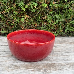 May include: A round, deep ceramic bowl with a glossy, vibrant red glaze. The bowl features a smooth, curved interior and exterior surface with subtle variations in the red colour intensity, creating a reflective finish on the rim and base.