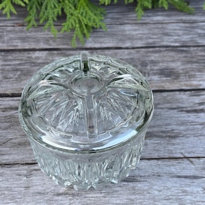 May include: A clear glass dish with a lid, featuring a cut-glass pattern. The round dish has a small handle on the lid. It sits on a weathered wooden surface, with green foliage in the background. A decorative storage container, ideal for small items.
