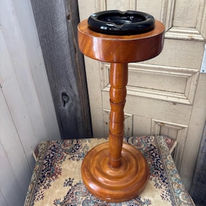 Könnte beinhalten: Ein Vintage-Aschenbecherständer aus Holz mit rundem Sockel und einem schwarzen Aschenbecher. Der Ständer ist aus poliertem Holz gefertigt und hat ein klassisches Design. Der Aschenbecher ist kreisförmig und sitzt oben auf dem Ständer. Der Ständer ist ca. 76 cm hoch.