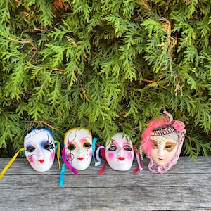 Peut inclure: Quatre masques décoratifs en céramique avec des visages peints et des rubans colorés. Les masques présentent différents motifs, avec des accents bleus, jaunes et roses. Ils sont disposés sur une surface en bois vieilli devant un fond de feuillage vert.