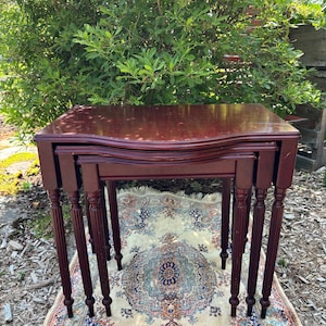 May include: A set of three nesting tables in a dark cherry finish. The tables feature a curved top and turned legs, displaying a classic design. The tables are placed on a patterned rug. The tables are a classic design.