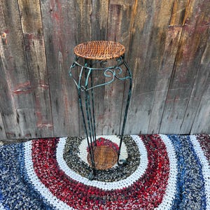 May include: A tall, decorative plant stand with a round, woven top and a wrought iron base. The metal base has a distressed, verdigris finish and features decorative scrollwork. The stand is approximately 76 cm tall.