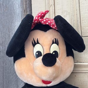 May include: A plush Minnie Mouse doll with black ears, a black nose, and a red bow with white polka dots. The doll has large, expressive eyes and a red mouth. The doll's body is black.