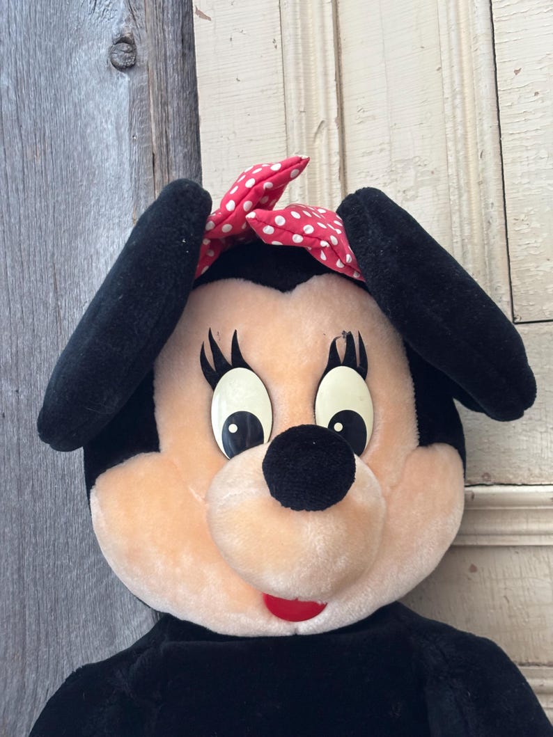May include: A plush Minnie Mouse doll with black ears, a black nose, and a red bow with white polka dots. The doll has large, expressive eyes and a red mouth. The doll's body is black.