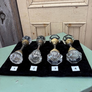 May include: Four vintage glass doorknobs with brass fittings are presented on a black surface. Each knob features a distinct faceted design and is numbered 1 to 4 on small white labels. A wooden door forms the backdrop.
