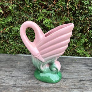 Könnte beinhalten: Eine Vintage-Schwanenvase aus Keramik in Pastelltönen. Die Vase hat einen rosa Körper mit einem flügelartigen Design und einen grünen Sockel. Der Schwanenhals bildet einen Griff. Die Vase steht vor einem Hintergrund aus grünem Laub.