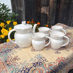 Puede incluir: Un juego de té de cerámica color crema que consta de una tetera con tapa y tres tazas a juego. Cada pieza presenta un patrón en relieve de uvas y hojas alrededor del borde superior y un motivo de hoja individual en la parte inferior.
