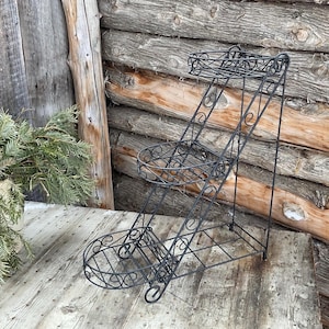May include: A black metal plant stand with three tiers. Each tier is a round, open-work design with a decorative scroll pattern. The stand is angled and has a rustic look.