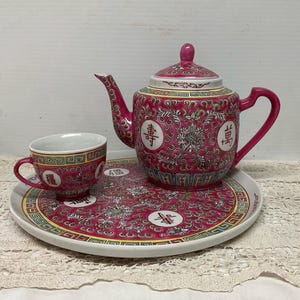 May include: A pink and white porcelain tea set with a teapot, cup, and saucer. The teapot and cup feature Chinese characters. The saucer has a floral design.