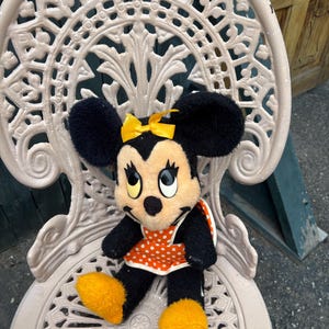 K&ouml;nnte beinhalten: Eine Minnie Mouse Pl&uuml;schpuppe mit schwarzem Fell, gro&szlig;en Ohren und einer gelben Schleife. Sie tr&auml;gt eine orangefarbene Sch&uuml;rze mit wei&szlig;en Punkten und gelbe Schuhe. Die Puppe sitzt auf einem wei&szlig;en, verzierten Metallstuhl.