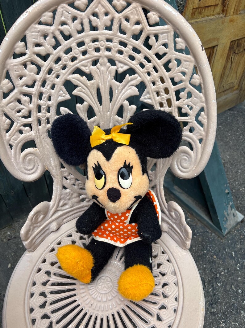 K&ouml;nnte beinhalten: Eine Minnie Mouse Pl&uuml;schpuppe mit schwarzem Fell, gro&szlig;en Ohren und einer gelben Schleife. Sie tr&auml;gt eine orangefarbene Sch&uuml;rze mit wei&szlig;en Punkten und gelbe Schuhe. Die Puppe sitzt auf einem wei&szlig;en, verzierten Metallstuhl.