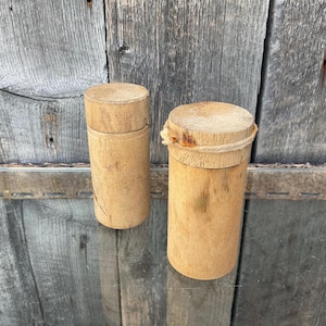 May include: Two cylindrical wooden containers with lids. One container has a rope tied around the lid. The wood has a light brown colour and a natural grain. The containers are set against a weathered wooden background.
