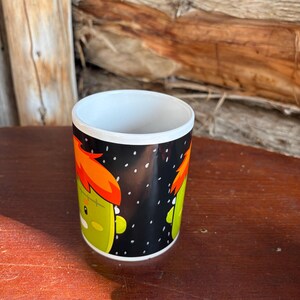 May include: A white ceramic mug featuring a black background with a repeating cartoon graphic of a green monster character with bright orange hair and a stitched forehead. The design includes small white dots scattered across the black surface.