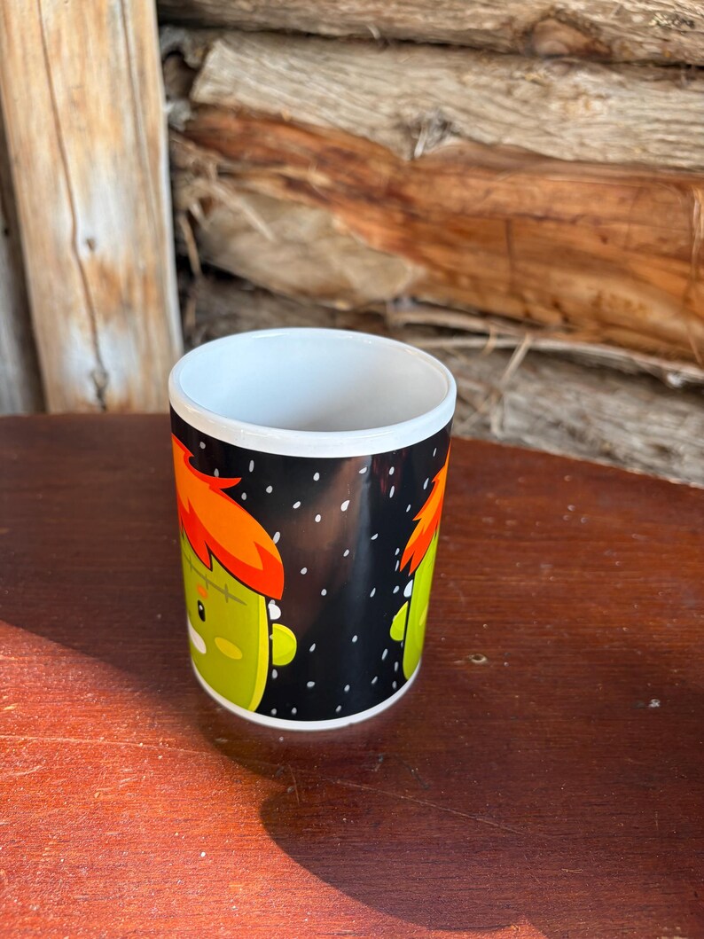 May include: A white ceramic mug featuring a black background with a repeating cartoon graphic of a green monster character with bright orange hair and a stitched forehead. The design includes small white dots scattered across the black surface.