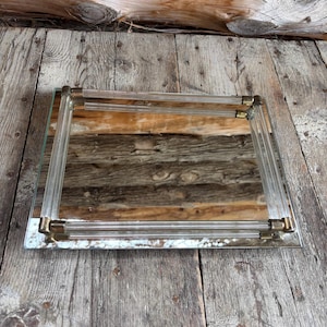 May include: Rectangular mirror with a clear acrylic frame and brass-coloured corner accents. The mirror's frame has a layered design, and the glass reflects the surrounding environment. The mirror is placed on a weathered wooden surface.