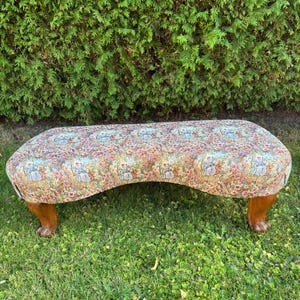 May include: An upholstered bench with a curved design and four wooden legs. The bench features a floral pattern in shades of pink, blue, and green. The bench is set on a grassy lawn with a green hedge in the background.