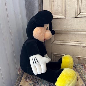May include: A large plush Mickey Mouse toy, primarily black with white gloves and yellow feet. The toy has a round, peach-colored face and a black hat. It is seated on a patterned rug.