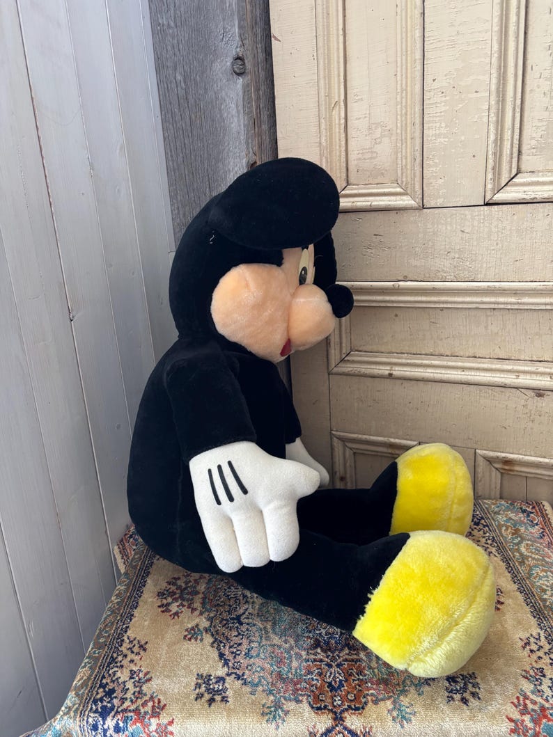 May include: A large plush Mickey Mouse toy, primarily black with white gloves and yellow feet. The toy has a round, peach-colored face and a black hat. It is seated on a patterned rug.