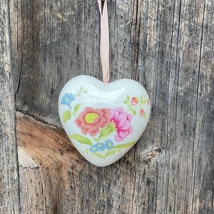 May include: A white porcelain heart-shaped ornament with a floral design. The flowers are pink, yellow, and blue. The ornament is hanging from a pink ribbon.