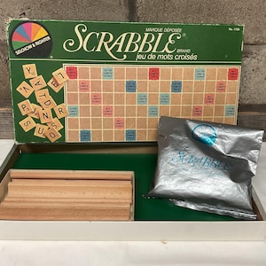 Pode incluir: Um jogo de tabuleiro Scrabble vintage numa caixa com um tabuleiro de jogo verde, peças de letras e um saco de peças. A caixa é vermelha, branca e azul com a palavra "Scrabble" em letras grandes.