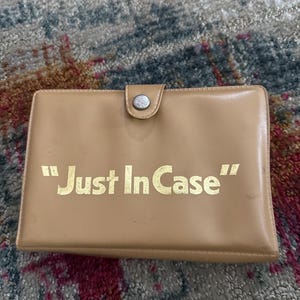 Könnte beinhalten: Eine braune Ledertasche mit einem silbernen Druckknopfverschluss. Die Tasche trägt den Schriftzug "Just In Case" in goldenen Buchstaben.