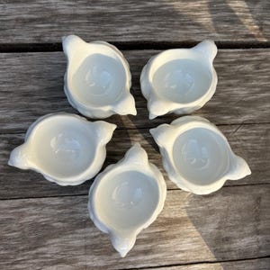 Puede incluir: Conjunto de cinco hueveras de cerámica blanca con forma de pollo. Cada huevera tiene un pequeño pico y dos pequeñas alas.