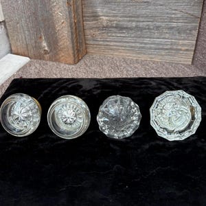 May include: Four clear glass doorknobs with detailed starburst and geometric patterns. The doorknobs are displayed on a black velvet surface, highlighting their reflective properties. The knobs have different shapes, some round and others faceted.